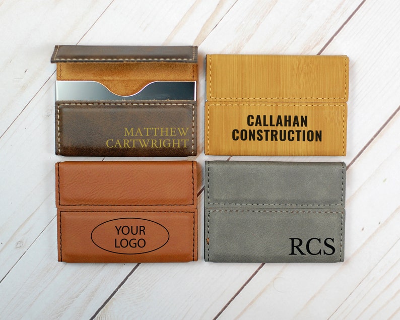 May include: Four different business card holders with various colors and materials. The first is brown leather with the name "Matthew Cartwright" in gold lettering. The second is a light brown wood with the text "CALLAHAN CONSTRUCTION" in black lettering. The third is a brown leather with the text "YOUR LOGO" in an oval shape. The fourth is a gray leather with the initials "RCS" in black lettering.