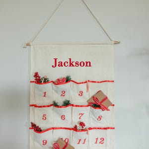 May include: A cream-colored Christmas advent calendar with red accents and the name "Jackson" printed at the top. The calendar has 12 pockets, each numbered and decorated with festive sprigs and small gifts.