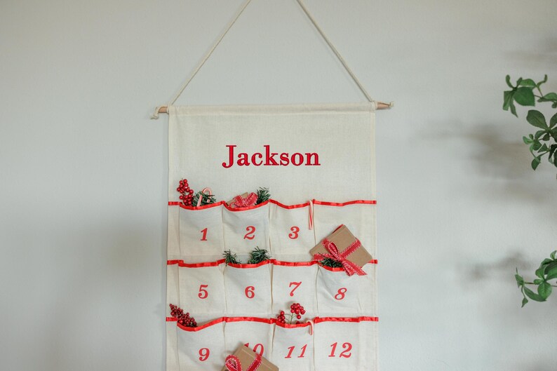 May include: A cream-colored Christmas advent calendar with red accents and the name "Jackson" printed at the top. The calendar has 12 pockets, each numbered and decorated with festive sprigs and small gifts.
