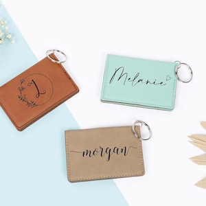 May include: Three personalized leather keychains in brown, mint green, and tan. The brown keychain features a letter "J" inside a wreath. The mint green keychain has the name "Melanie" written on it. The tan keychain has the name "morgan" written on it.