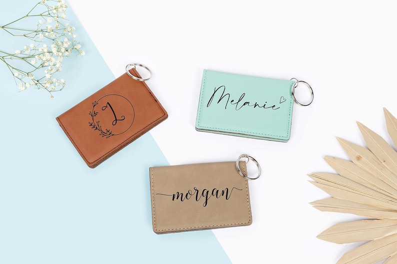 May include: Three personalized leather keychains in brown, mint green, and tan. The brown keychain features a letter "J" inside a wreath. The mint green keychain has the name "Melanie" written on it. The tan keychain has the name "morgan" written on it.