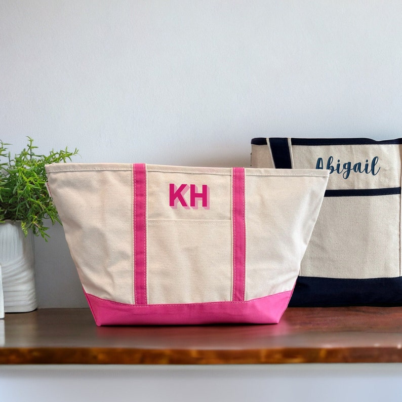 May include: Two canvas tote bags with pink trim. The bag on the left has the initials "KH" embroidered on the front in pink. The bag on the right has the name "Abigail" embroidered on the front in navy blue.
