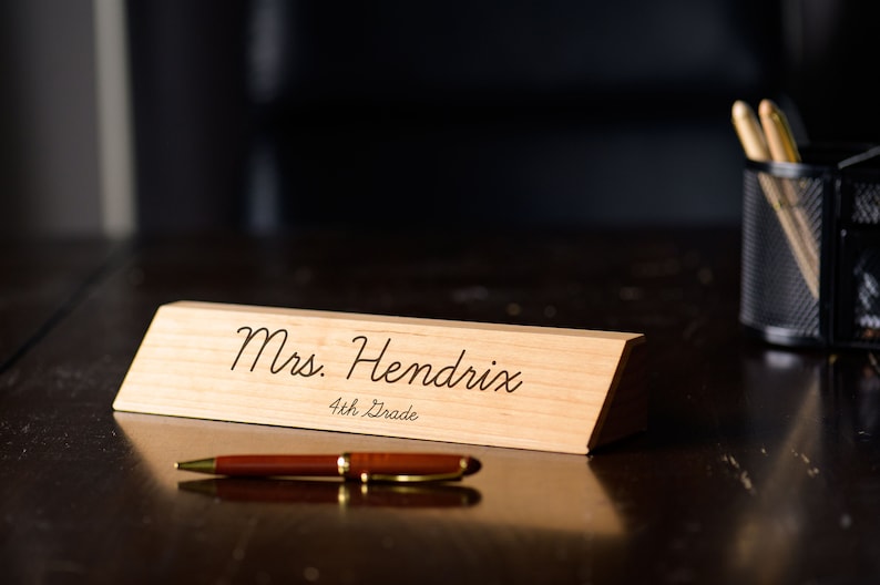 May include: A wooden desk nameplate with the text "Mrs. Hendrix 4th Grade" engraved on it. The nameplate is sitting on a dark wooden desk.