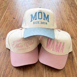 May include: Three baseball caps with embroidered text. One cap is cream-coloured with a light blue brim and the word "MOM EST.2026" in blue. The other two caps are cream-coloured with pink brims, one reading "OVERSTIMULATED Mom Club JAMES 1:2-4" and the other "MAMA".