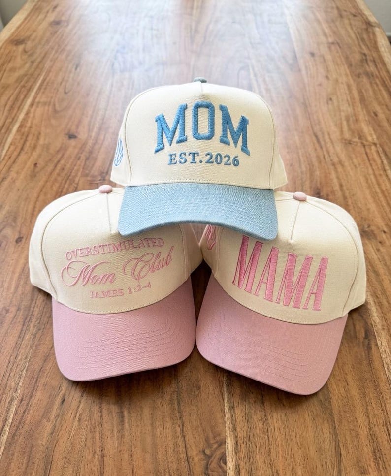 May include: Three baseball caps with embroidered text. One cap is cream-coloured with a light blue brim and the word "MOM EST.2026" in blue. The other two caps are cream-coloured with pink brims, one reading "OVERSTIMULATED Mom Club JAMES 1:2-4" and the other "MAMA".