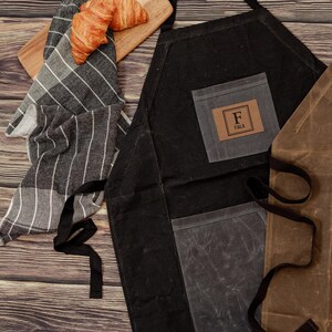 May include: Black canvas apron with a large pocket and a smaller pocket with a brown leather patch that says "F  FIALA". The apron is on a wooden surface with a striped kitchen towel and a croissant.