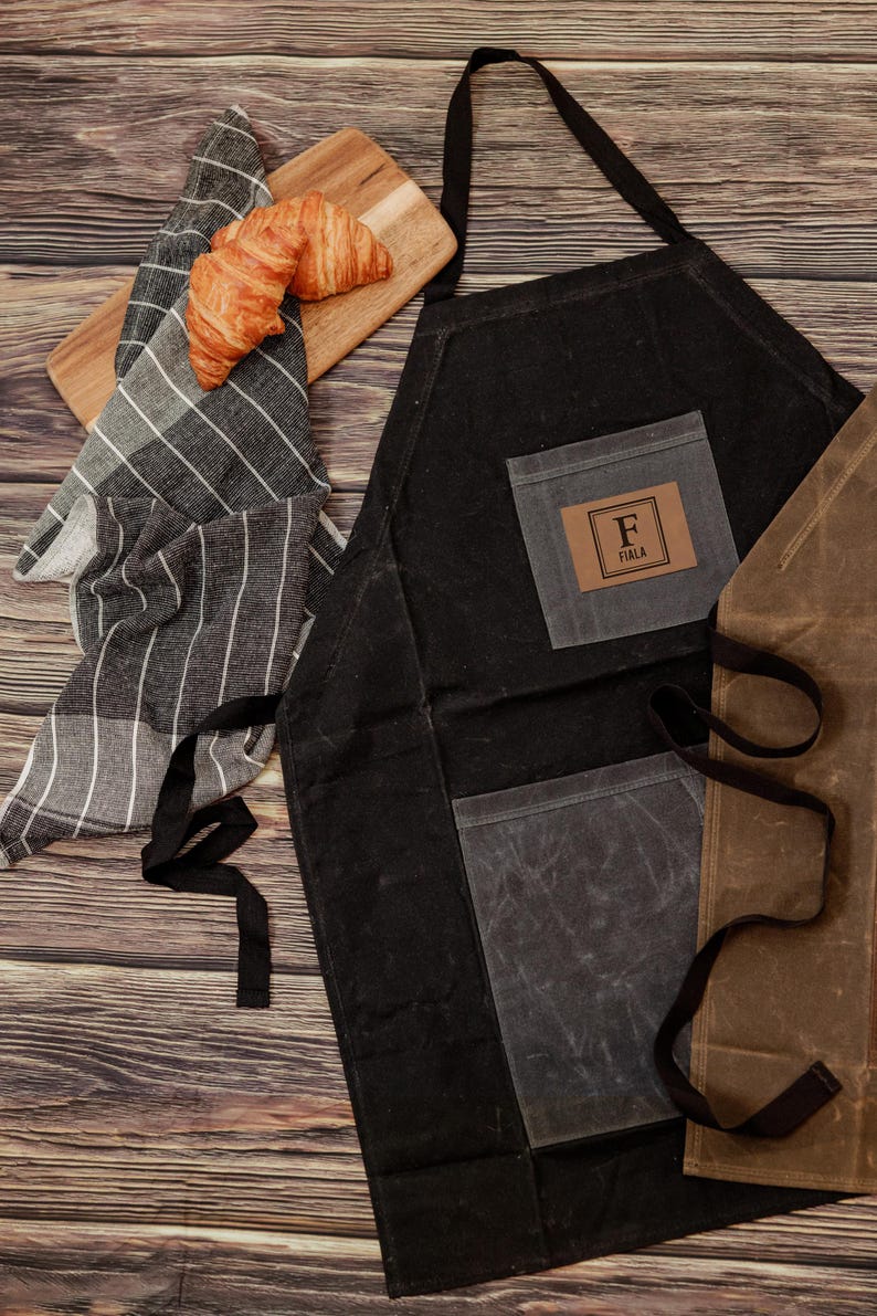 May include: Black canvas apron with a large pocket and a smaller pocket with a brown leather patch that says "F  FIALA". The apron is on a wooden surface with a striped kitchen towel and a croissant.