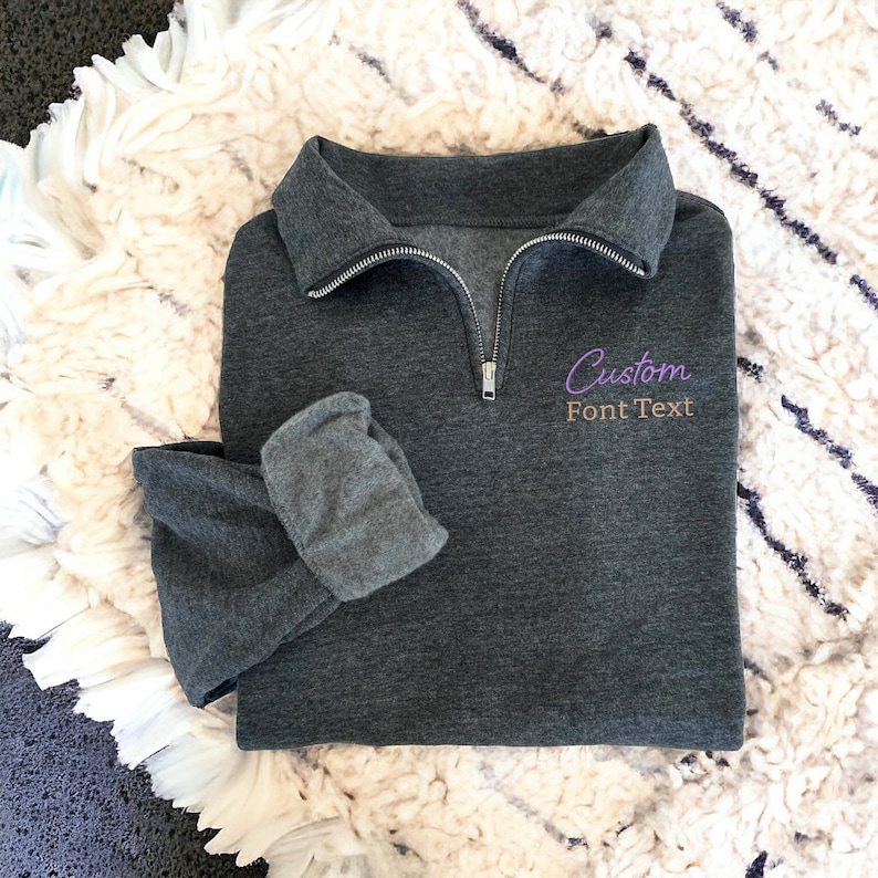 May include: A dark grey quarter-zip sweatshirt with a purple embroidered design that says "Custom Font Text".