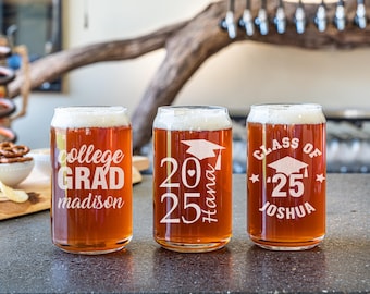 May include: Three clear glass beer can-shaped glasses filled with beer and foam. Each glass has white text: "college GRAD madison", "2025 Hana", and "CLASS OF '25 joshua". The glasses are on a dark surface.