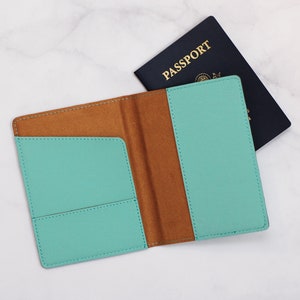 May include: A teal passport holder with a brown interior and a stitched edge. The holder is open and the inside is visible. The holder is on a white marble surface.