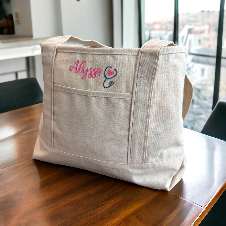 May include: A beige canvas tote bag with a pink embroidered stethoscope and the name "Alyssa" on the front.
