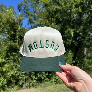 May include: A cream-colored baseball cap with a dark green brim and the word "CUSTOM" embroidered in green. The hat is held up against a backdrop of green trees and a blue sky.
