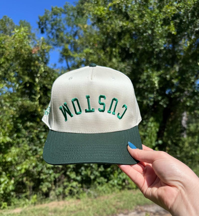 May include: A cream-colored baseball cap with a dark green brim and the word "CUSTOM" embroidered in green. The hat is held up against a backdrop of green trees and a blue sky.