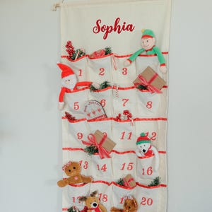 May include: Cream-colored fabric advent calendar with 24 pockets, red trim, and the name "Sophia" in red script. Each pocket contains a small toy or decoration, including a Santa, elf, bear, and reindeer. Numbers 1-24 are printed in red.