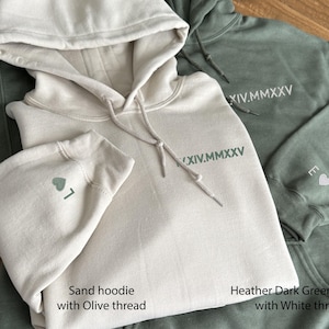 May include: Three hooded sweatshirts in sand and heather dark green. The sand hoodie has olive thread embroidery, including a heart and the number 7 on the sleeve. The dark green hoodie has white thread embroidery with the roman numerals XIV.MMXXV.