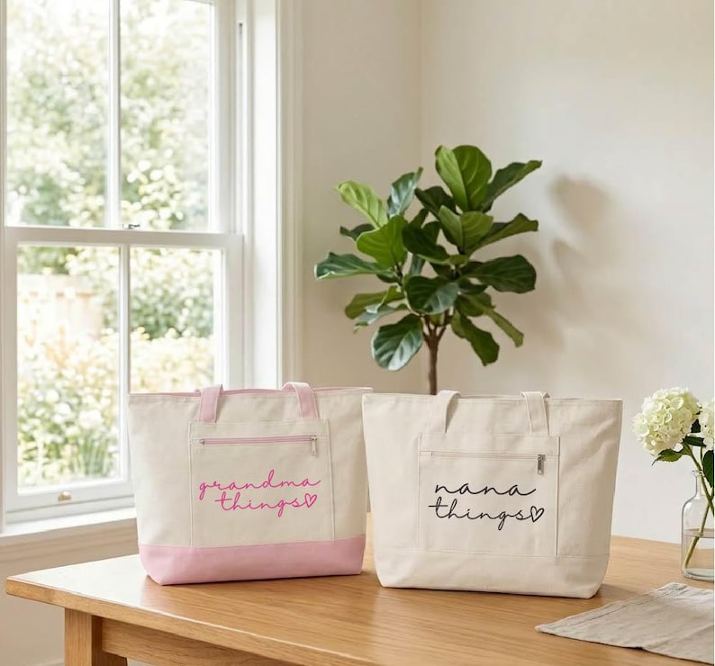 Op de afbeelding: Twee canvas draagtassen met ritsvakken aan de voorkant. E&eacute;n is gebroken wit met een roze bodem en handvatten, en de woorden "grandma things" in roze script. De andere is gebroken wit met de woorden "nana things" in zwart script.