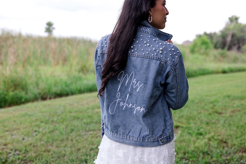 May include: A blue denim jacket with pearl embellishments on the shoulders and the text "Mrs. Johnson" embroidered on the back in white.