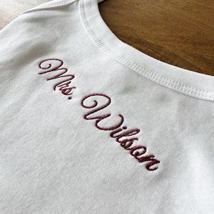May include: White tank top with burgundy embroidered text reading "Mrs. Wilson". The top features a scoop neckline and thin straps. The embroidery is in a cursive font.