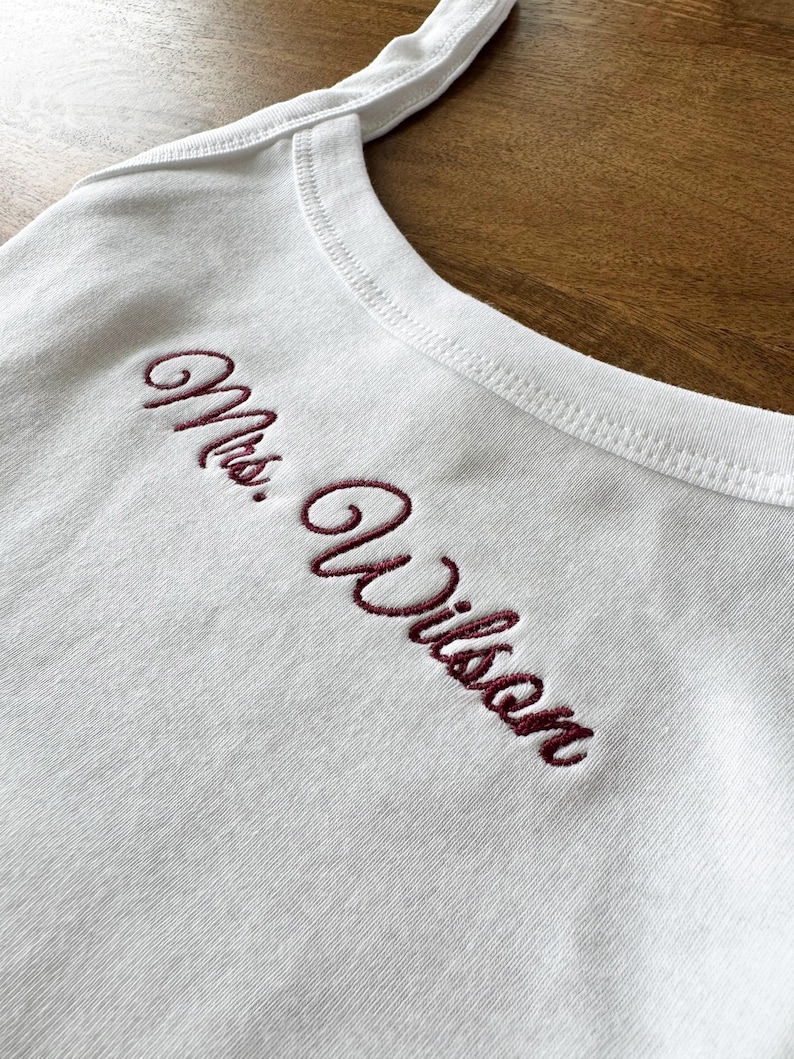 May include: White tank top with burgundy embroidered text reading "Mrs. Wilson". The top features a scoop neckline and thin straps. The embroidery is in a cursive font.