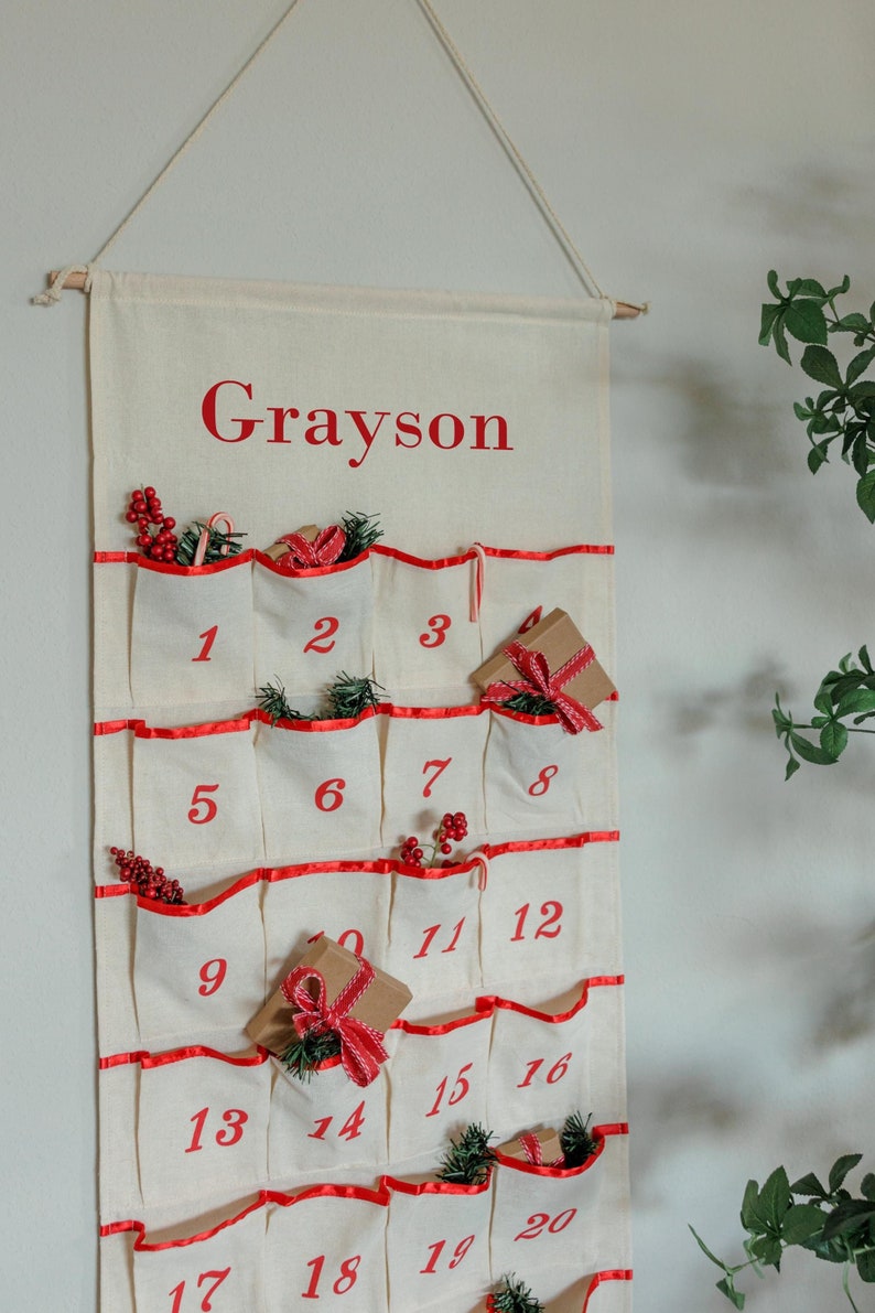 May include: Cream-colored fabric advent calendar with red trim and pockets numbered 1-20. The name "Grayson" is printed in red at the top. Some pockets contain small gifts, candy canes, and sprigs of greenery. A wooden dowel and rope are used for hanging.
