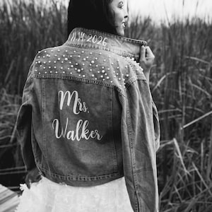 May include: A black and white photo of a person wearing a denim jacket with pearl embellishments. The jacket has the text "Mrs. Walker" and the date "11.14.2026" written on the back.