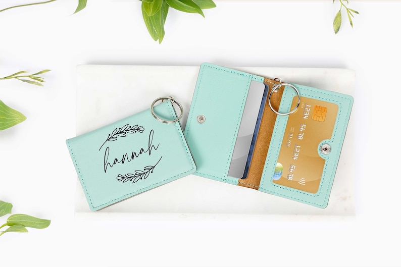 May include: A light teal leather cardholder and key ring set. One cardholder is open, displaying a credit card and a silver card. The other cardholder is closed and has the name "hannah" printed on it with a floral design.
