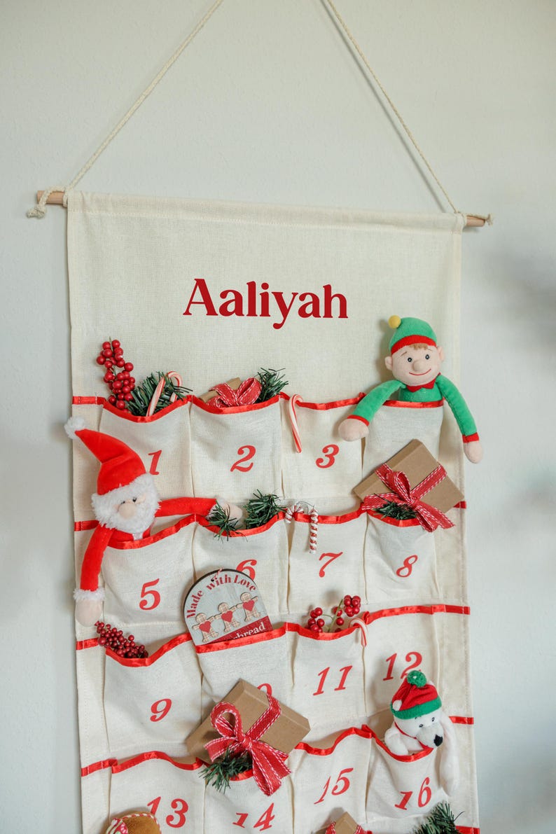 May include: A cream-colored advent calendar with red trim and pockets numbered 1-16. The name "Aaliyah" is printed in red at the top. The pockets contain small gifts and festive decorations, including a Santa, an elf, and a polar bear.