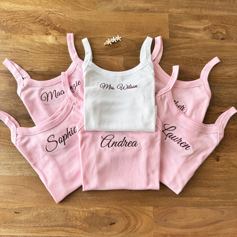 May include: A collection of pink and white tank tops with embroidered names and the text "Mrs. Wilson". The tank tops are arranged on a wooden surface, with a small floral accent.