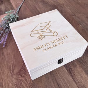 May include: A wooden box with a hinged lid and a clasp. The box is engraved with a graduation cap and scroll, and the text "ASHLEY NESBITT CLASS OF 2025".