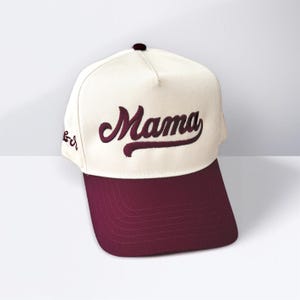 May include: A baseball cap with a cream-coloured crown and a maroon brim. The word "Mama" is embroidered in maroon script across the front. The cap has a curved brim and a button on top.