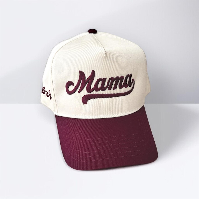 May include: A baseball cap with a cream-coloured crown and a maroon brim. The word "Mama" is embroidered in maroon script across the front. The cap has a curved brim and a button on top.