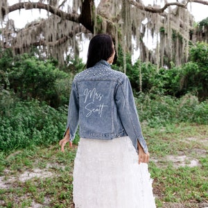 May include: A woman wearing a white lace skirt and a blue denim jacket with pearl embellishments. The back of the jacket has the text "Mrs. Scott" written in white.
