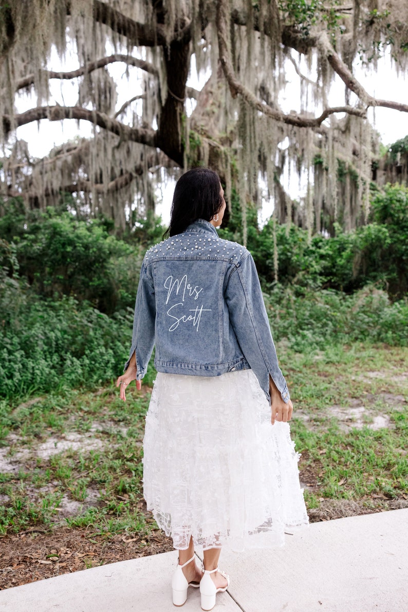May include: A woman wearing a white lace skirt and a blue denim jacket with pearl embellishments. The back of the jacket has the text "Mrs. Scott" written in white.