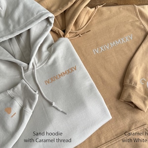 May include: Close-up of two hoodies, one sand-colored and one caramel-colored. Both have embroidered Roman numerals on the chest. The sand hoodie has caramel-colored thread, and the caramel hoodie has white thread. The sand hoodie has a small embroidered design on the sleeve.