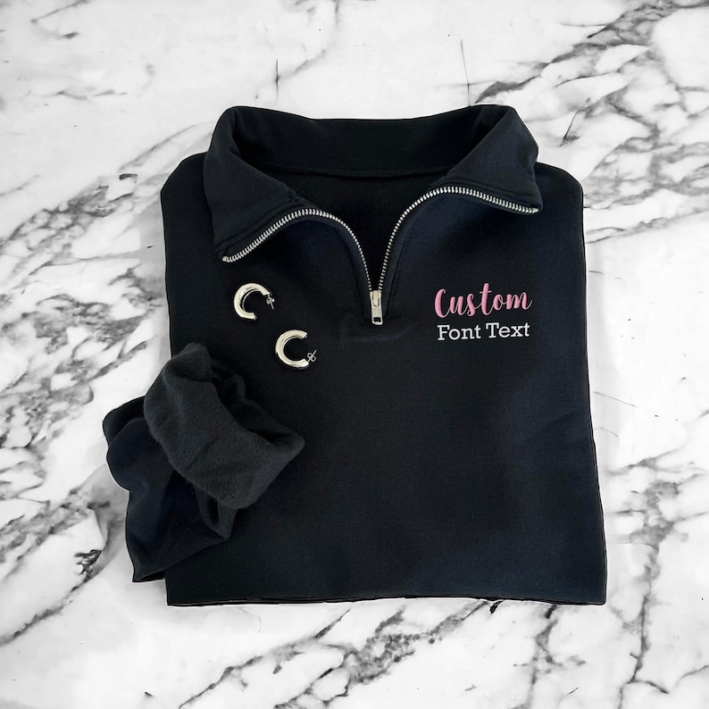 May include: Black quarter-zip sweatshirt with a pink embroidered design that says "Custom Font Text".