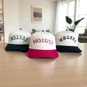 May include: Three white baseball caps with the word "CUSTOM" embroidered on the front. Each cap has a different colored brim: dark green, hot pink, and navy blue. The caps are displayed on a light brown surface.