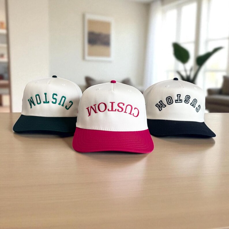 May include: Three white baseball caps with the word "CUSTOM" embroidered on the front. Each cap has a different colored brim: dark green, hot pink, and navy blue. The caps are displayed on a light brown surface.