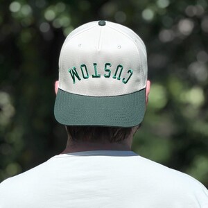 May include: A white and green baseball cap with the word "CUSTOM" embroidered in green across the front. The cap has a green brim and a dark green button on top. The person wearing the cap is facing away from the camera.