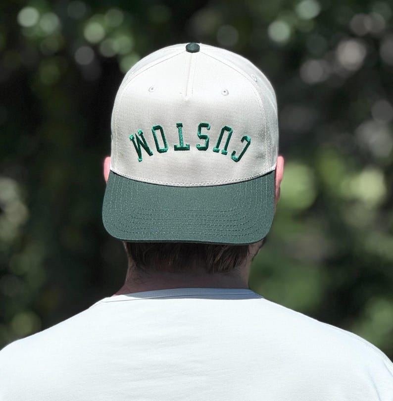 May include: A white and green baseball cap with the word "CUSTOM" embroidered in green across the front. The cap has a green brim and a dark green button on top. The person wearing the cap is facing away from the camera.