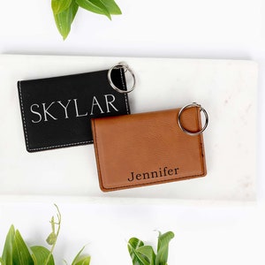 May include: Two personalized leather keychains, one black with "Skylar" and one brown with "Jennifer" engraved. Both have silver key rings and are displayed on a white marble tray with green foliage.