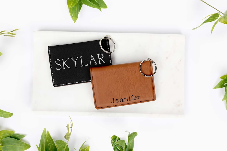 May include: Two personalized leather keychains, one black with "Skylar" and one brown with "Jennifer" engraved. Both have silver key rings and are displayed on a white marble tray with green foliage.
