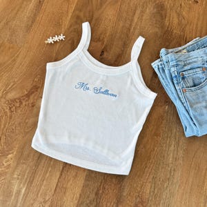 May include: White tank top with spaghetti straps and the embroidered name "Ms. Sullivan" in blue script. The top is paired with a pair of blue jeans. A gold hair clip with white flowers is also visible.