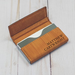 May include: A brown leather business card holder with a silver metal insert. The holder has the name "MATTHEW CARTWRIGHT" engraved on the front.