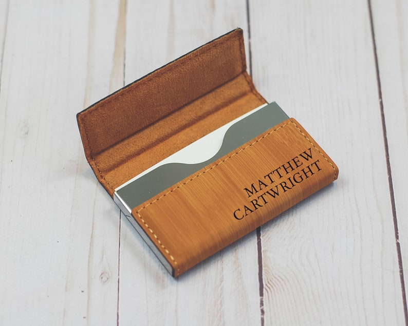 May include: A brown leather business card holder with a silver metal insert. The holder has the name "MATTHEW CARTWRIGHT" engraved on the front.