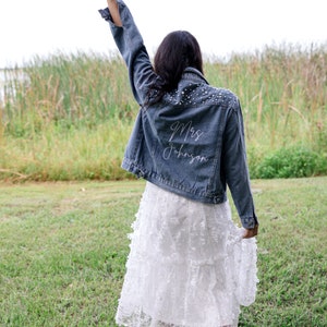 May include: A woman wearing a white lace dress and a denim jacket with pearl embellishments. The back of the jacket is embroidered with the text "Mrs. Johnson".