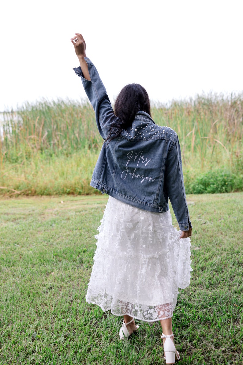 May include: A woman wearing a white lace dress and a denim jacket with pearl embellishments. The back of the jacket is embroidered with the text "Mrs. Johnson".