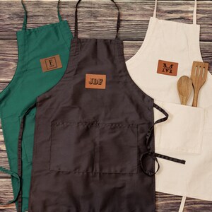 May include: Three personalized aprons with brown leather patches. The aprons are green, black, and beige. The patches have the initials "F", "JDF", and "M" on them.