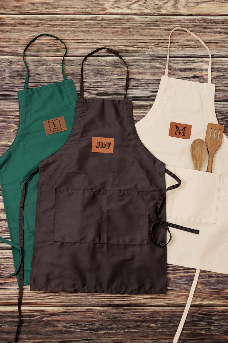 May include: Three personalized aprons with brown leather patches. The aprons are green, black, and beige. The patches have the initials "F", "JDF", and "M" on them.