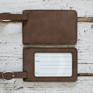 May include: Two brown leather luggage tags with a silver buckle. The top tag is blank, and the bottom tag has a white label with the words "Name", "Address", and "Phone" printed on it.