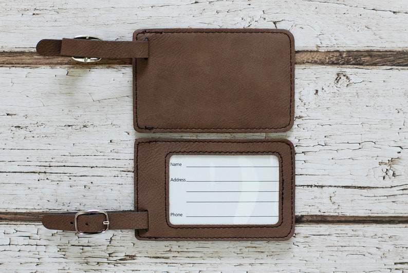 May include: Two brown leather luggage tags with a silver buckle. The top tag is blank, and the bottom tag has a white label with the words "Name", "Address", and "Phone" printed on it.